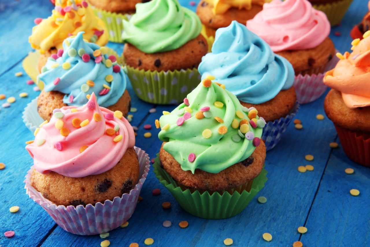 Um monte de cupcakes com cobertura e granulados de cores diferentes em uma mesa azul.