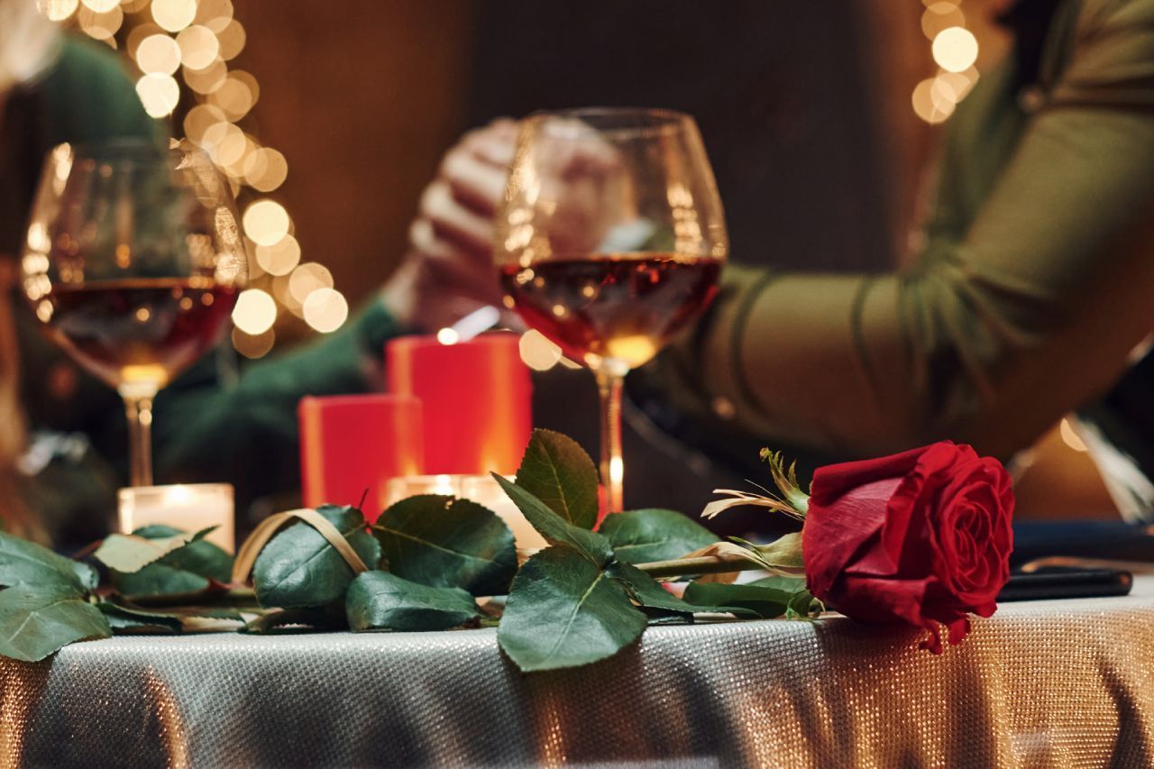 Uma mulher está sentada à mesa com uma rosa vermelha e taças de vinho.
