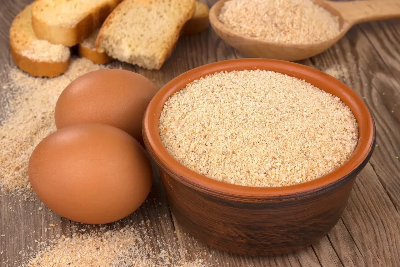 Uma tigela de farinha ao lado de ovos e pão em uma mesa de madeira.