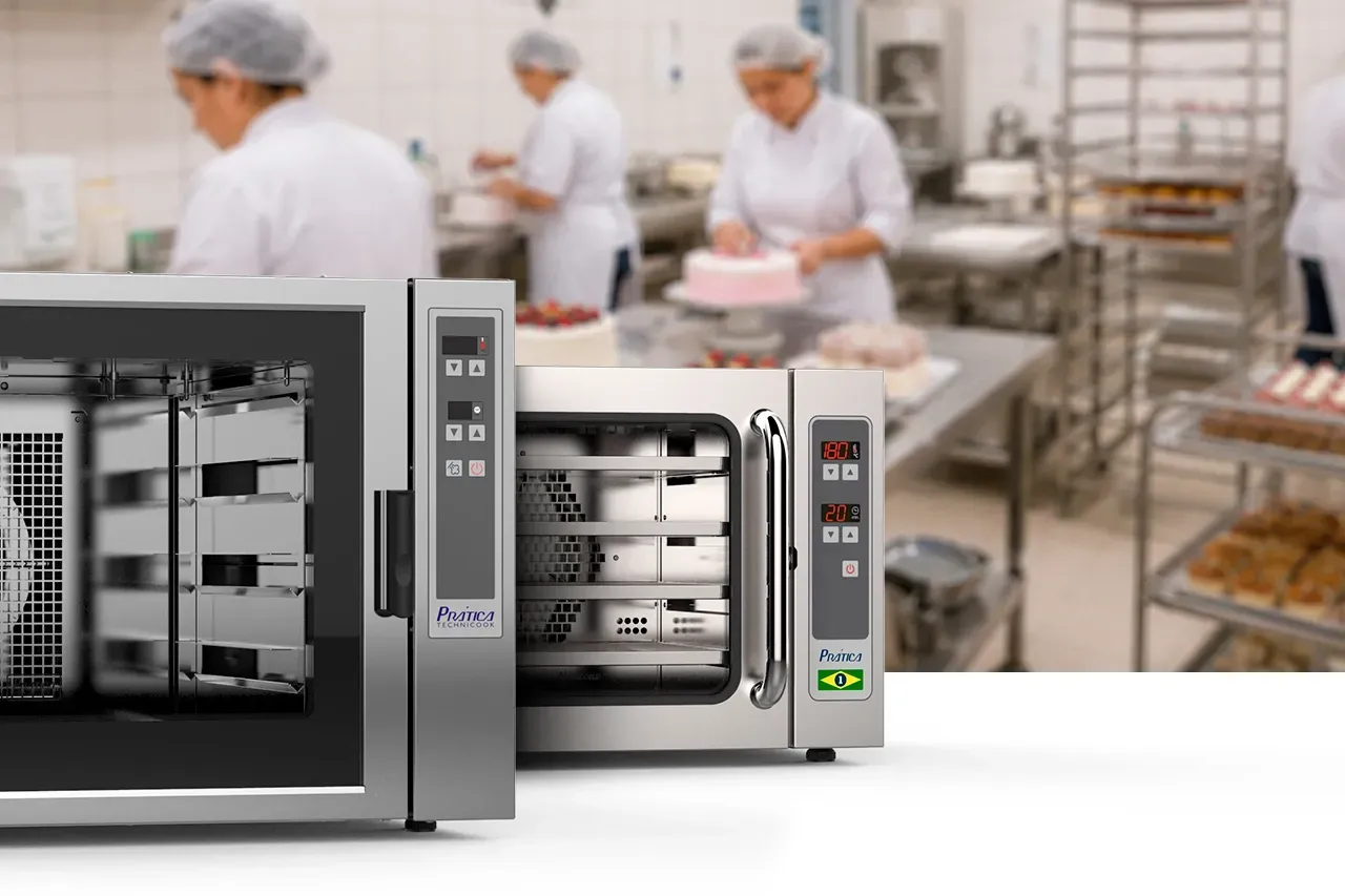 Fornos industriais Prática HPE80 e Miniconv em aço inox dispostos em primeiro plano. Ao fundo, cozinha profissional de confeitaria com funcionárias preparando bolos