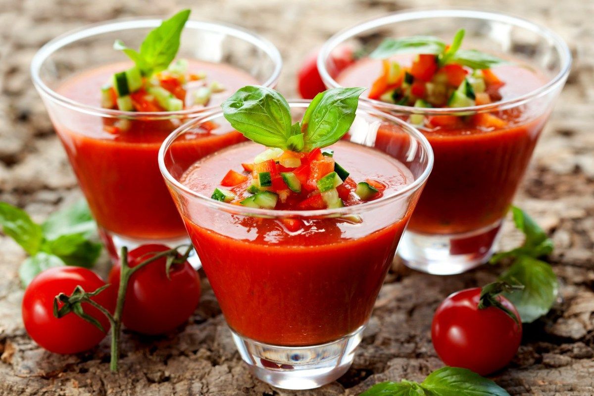 Três copos de gaspacho com tomates e pepinos sobre uma mesa de madeira.