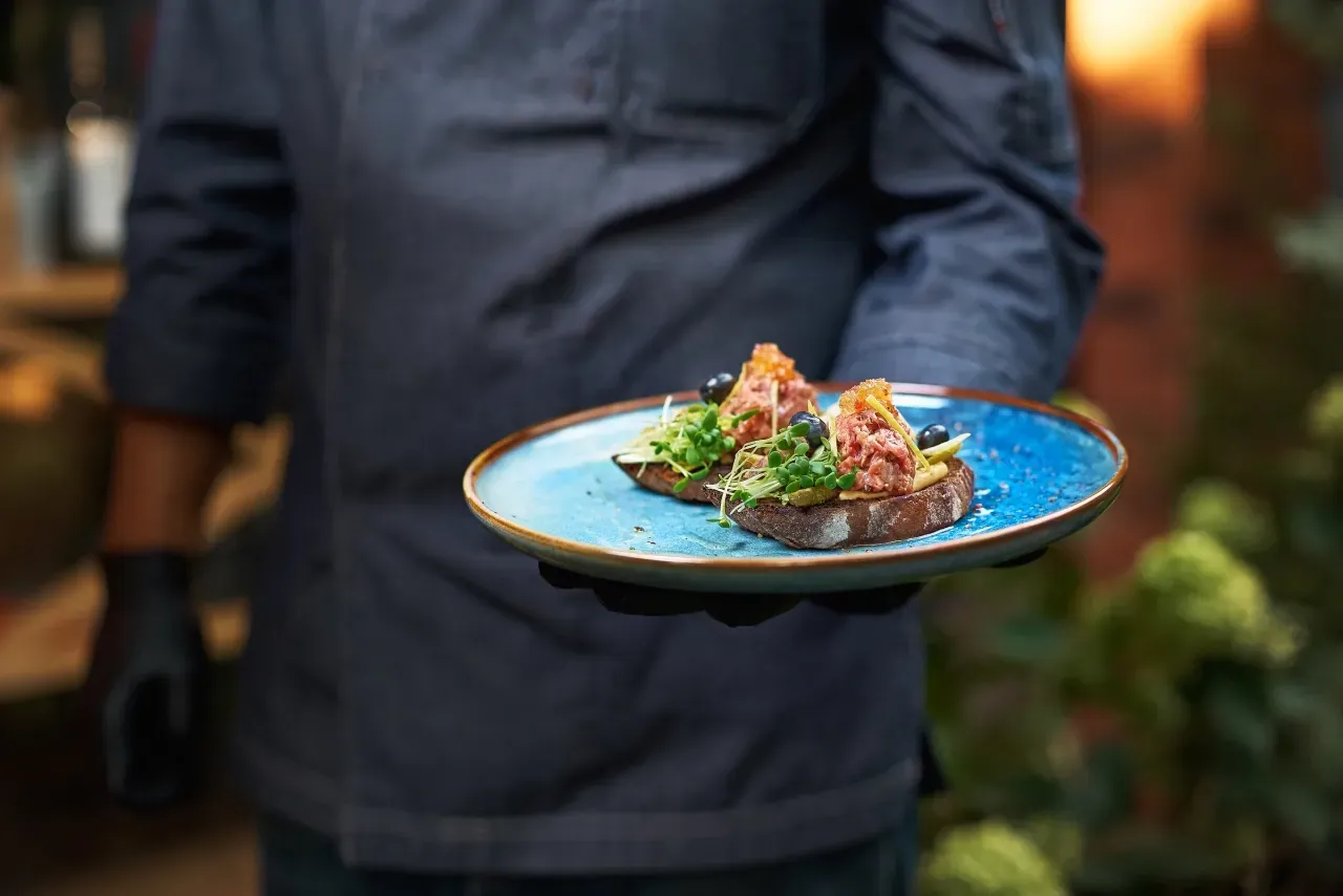 Chef segurando prato azul com duas torradas gourmet decoradas com brotos e guarnições frescas.