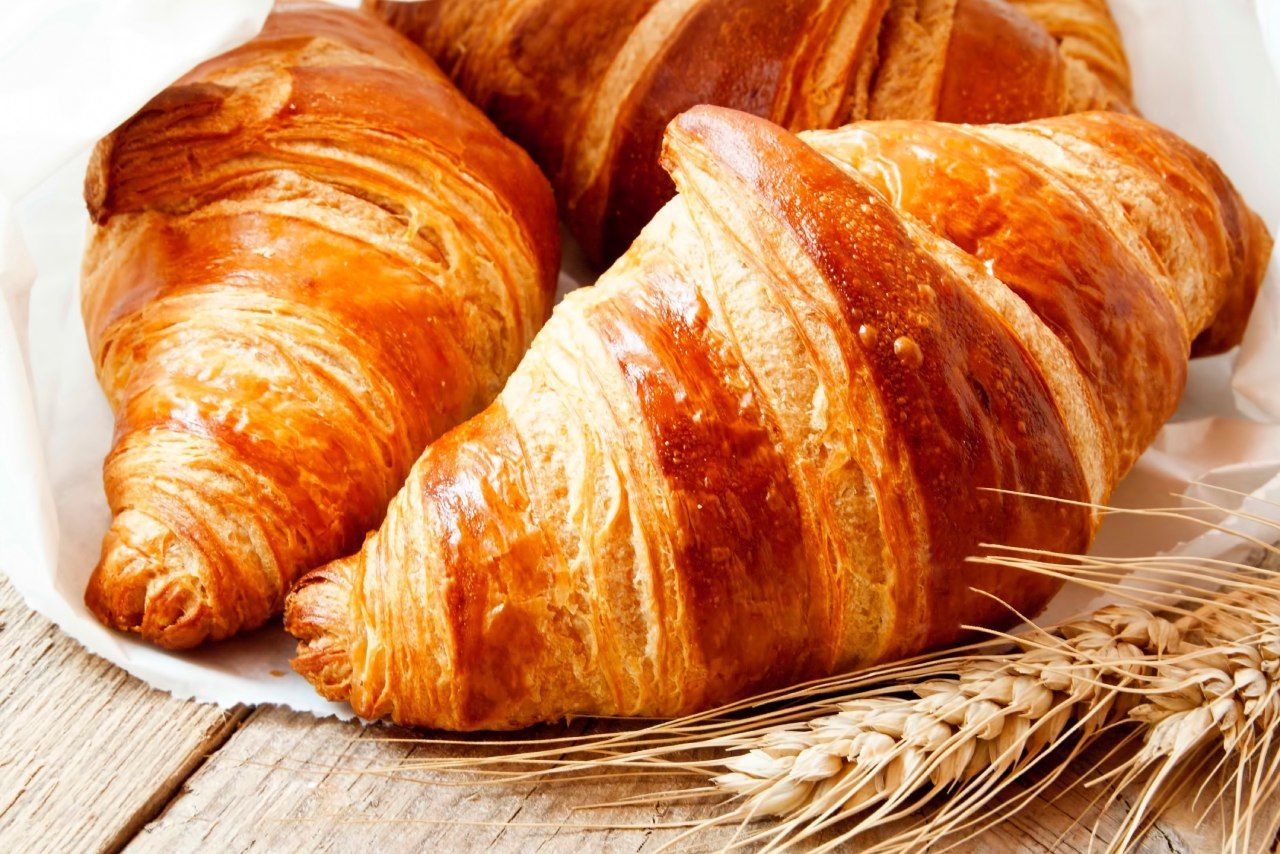 Três croissants estão sobre uma mesa de madeira ao lado de espigas de trigo.