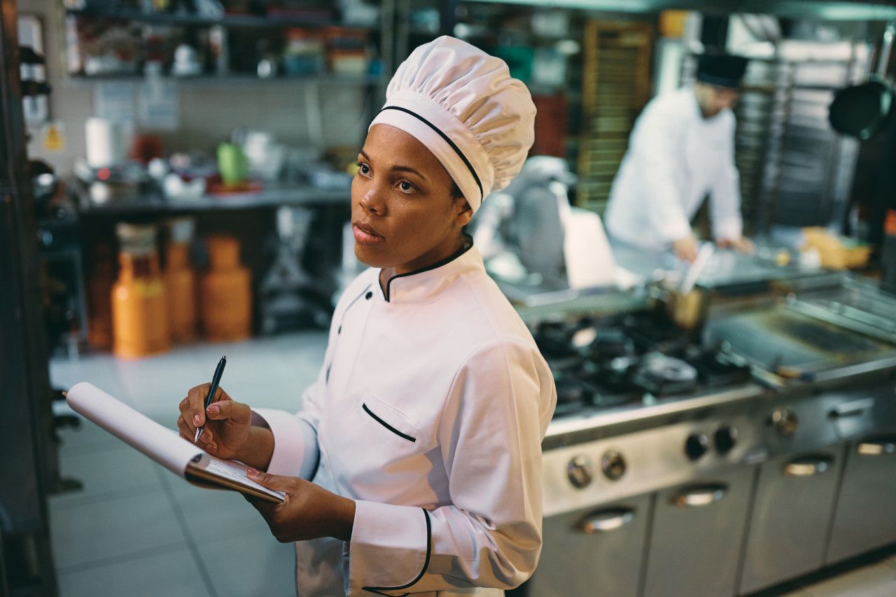 Uma mulher com um chapéu de chef está escrevendo em uma prancheta na cozinha.