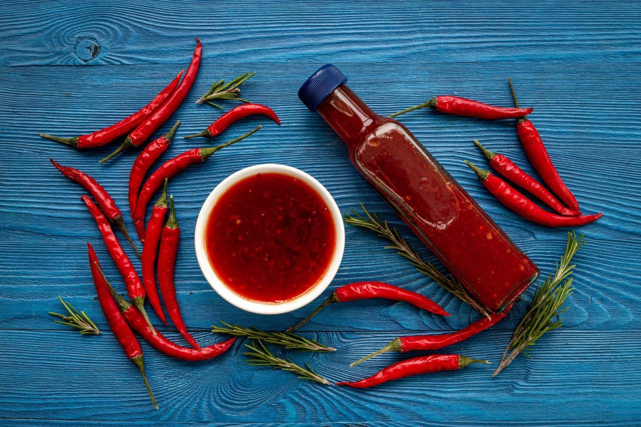 Uma garrafa de molho picante cercada por pimentões vermelhos sobre uma mesa de madeira azul.