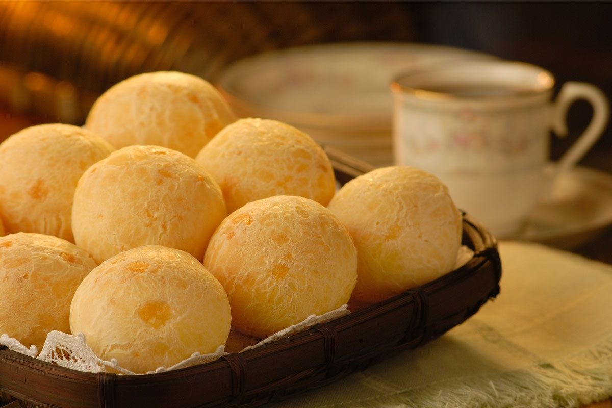 Uma cesta cheia pães de de queijo ao lado de uma xícara de café.