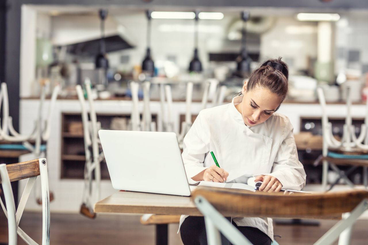 Uma mulher está sentada à mesa com um laptop e escrevendo em um caderno.
