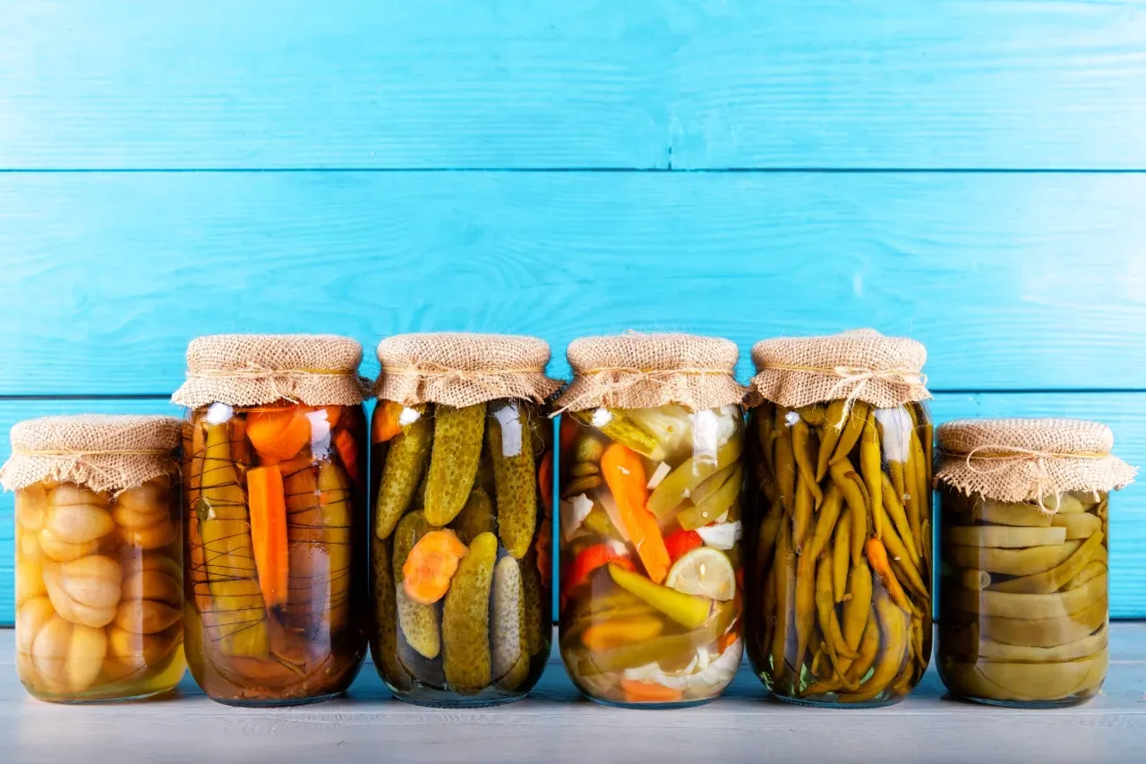 Potes de vidro com picles variados, como pepino, cenoura, cebola e vagem, conservados em líquido e alinhados sobre uma superfície de madeira azul.