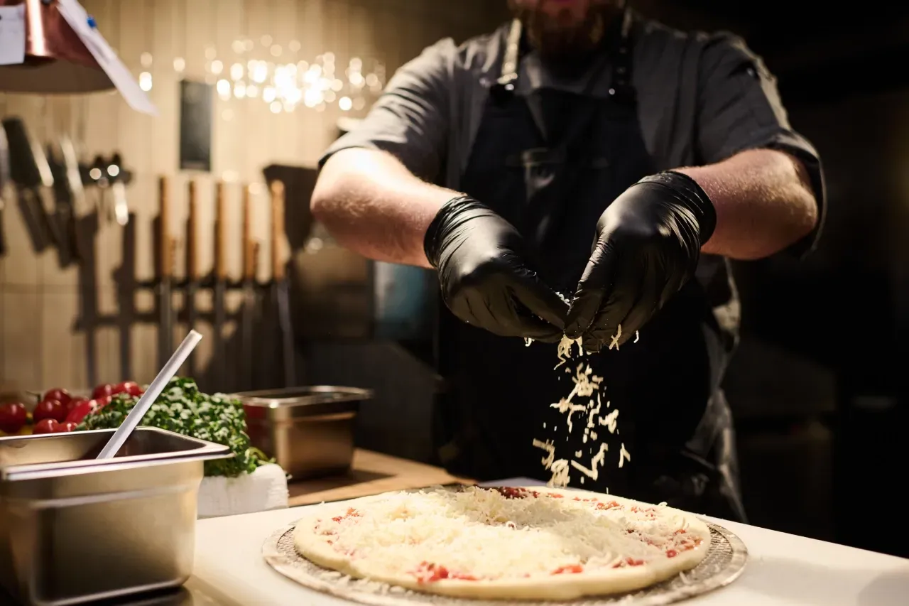 Um homem usando luvas pretas está espalhando queijo em uma pizza.
