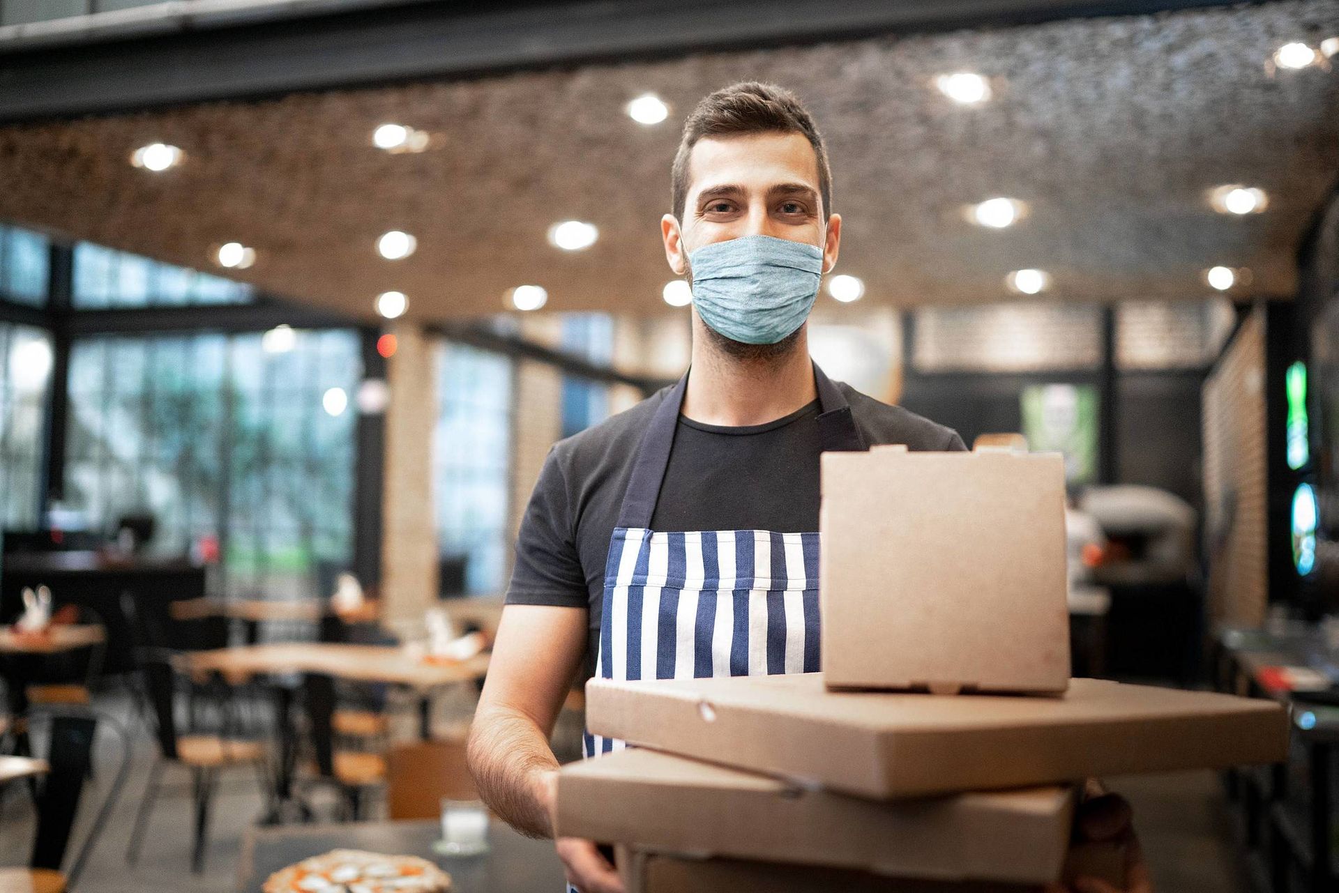 Um homem usando uma máscara está segurando uma pilha de caixas de pizza em um restaurante.