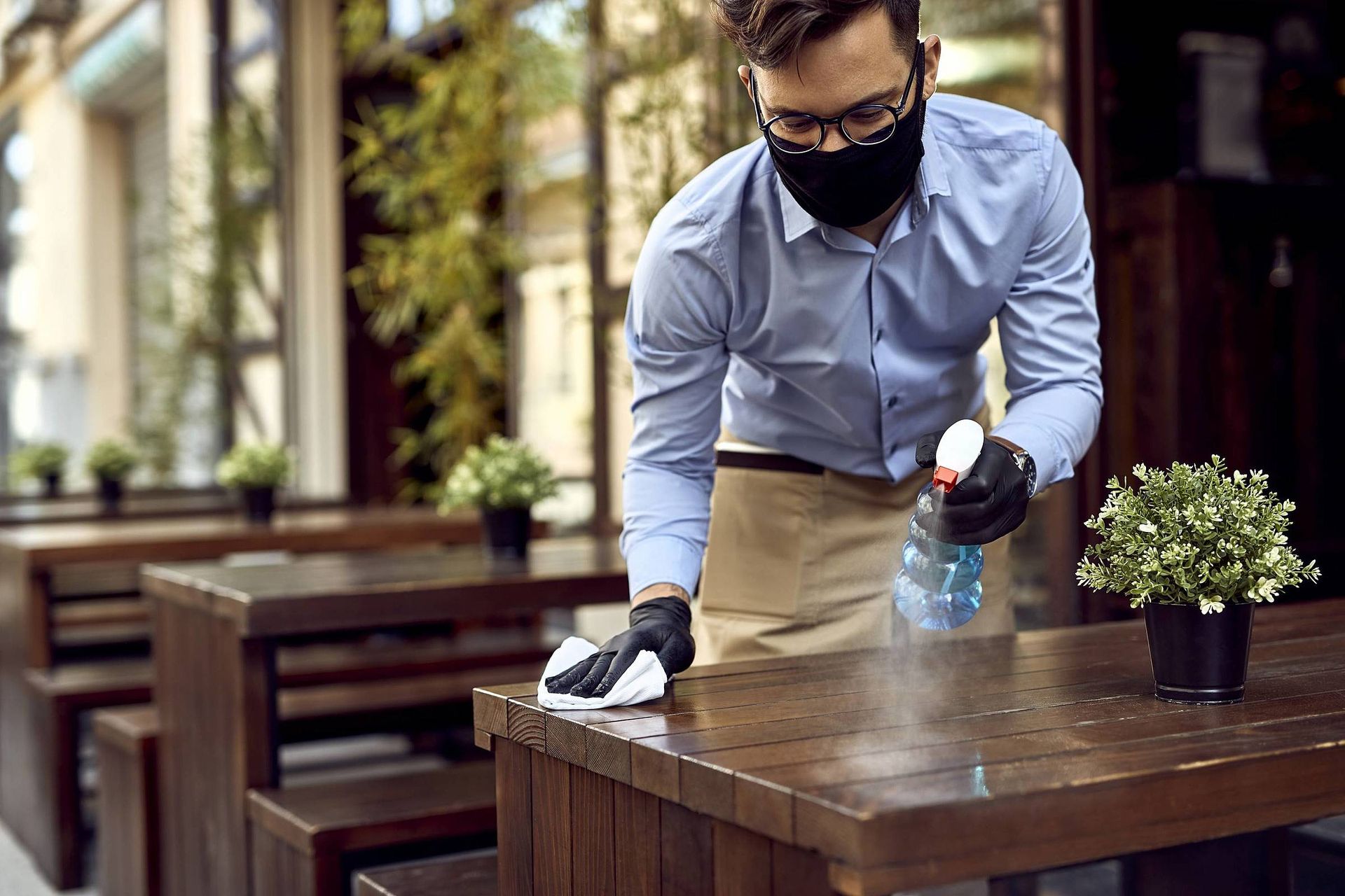 Um homem usando máscara e luvas está limpando uma mesa de madeira.