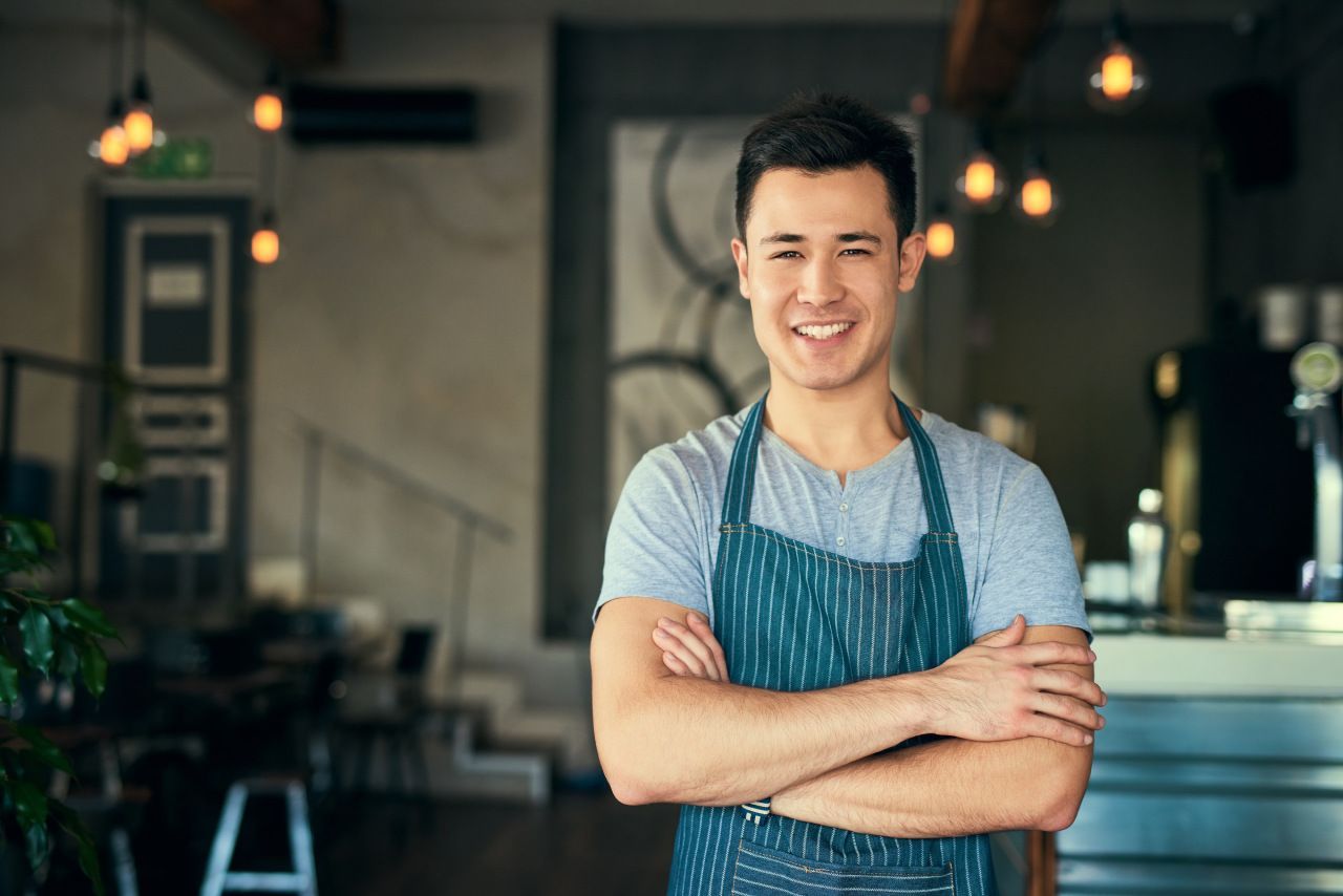 Um homem de avental está de pé em um restaurante com os braços cruzados.