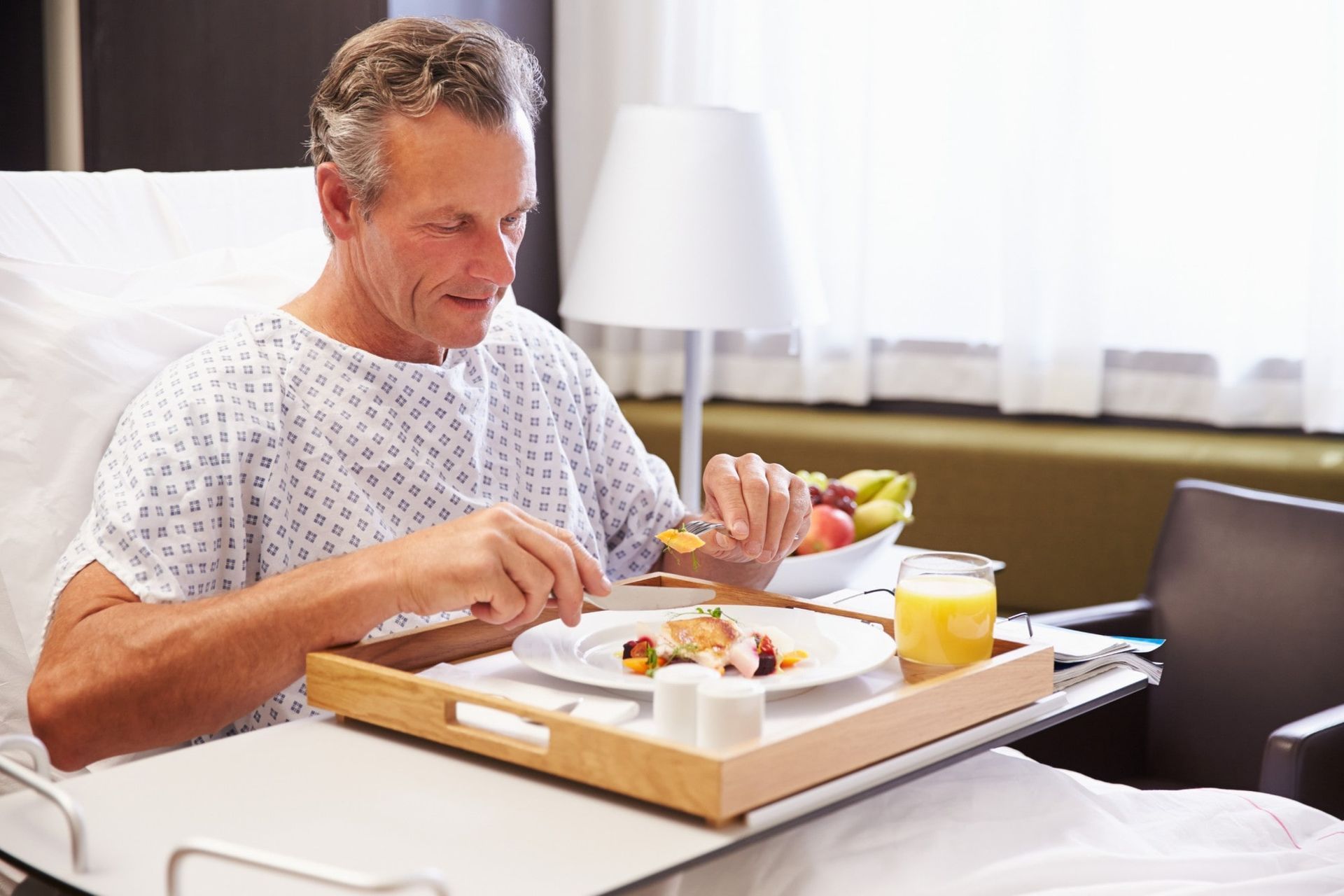Um homem está sentado em uma cama de hospital comendo comida de uma bandeja.