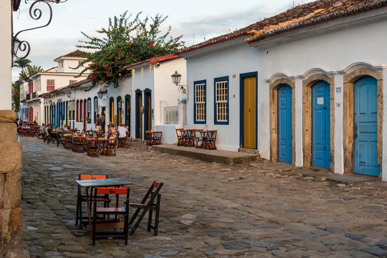 Uma fileira de edifícios com portas azuis, mesas e cadeiras em uma rua de paralelepípedos.