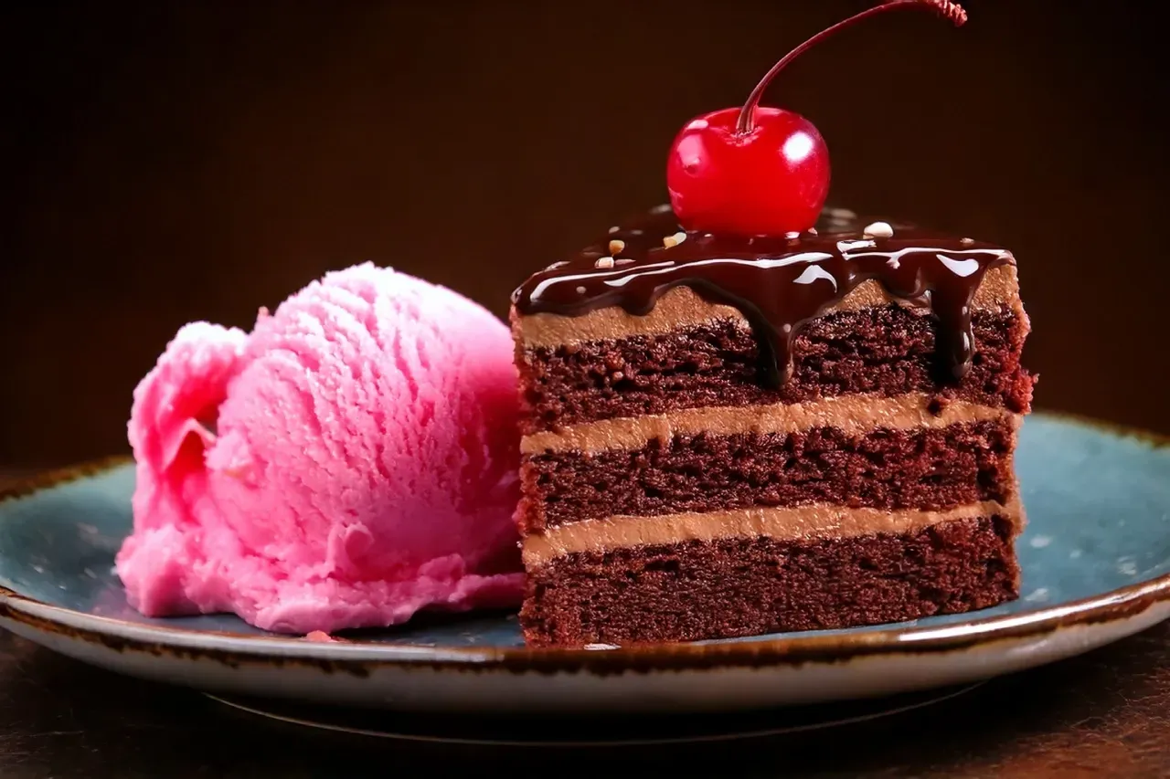 Um pedaço de bolo de chocolate com uma cereja no topo e uma bola de sorvete rosa em um prato.