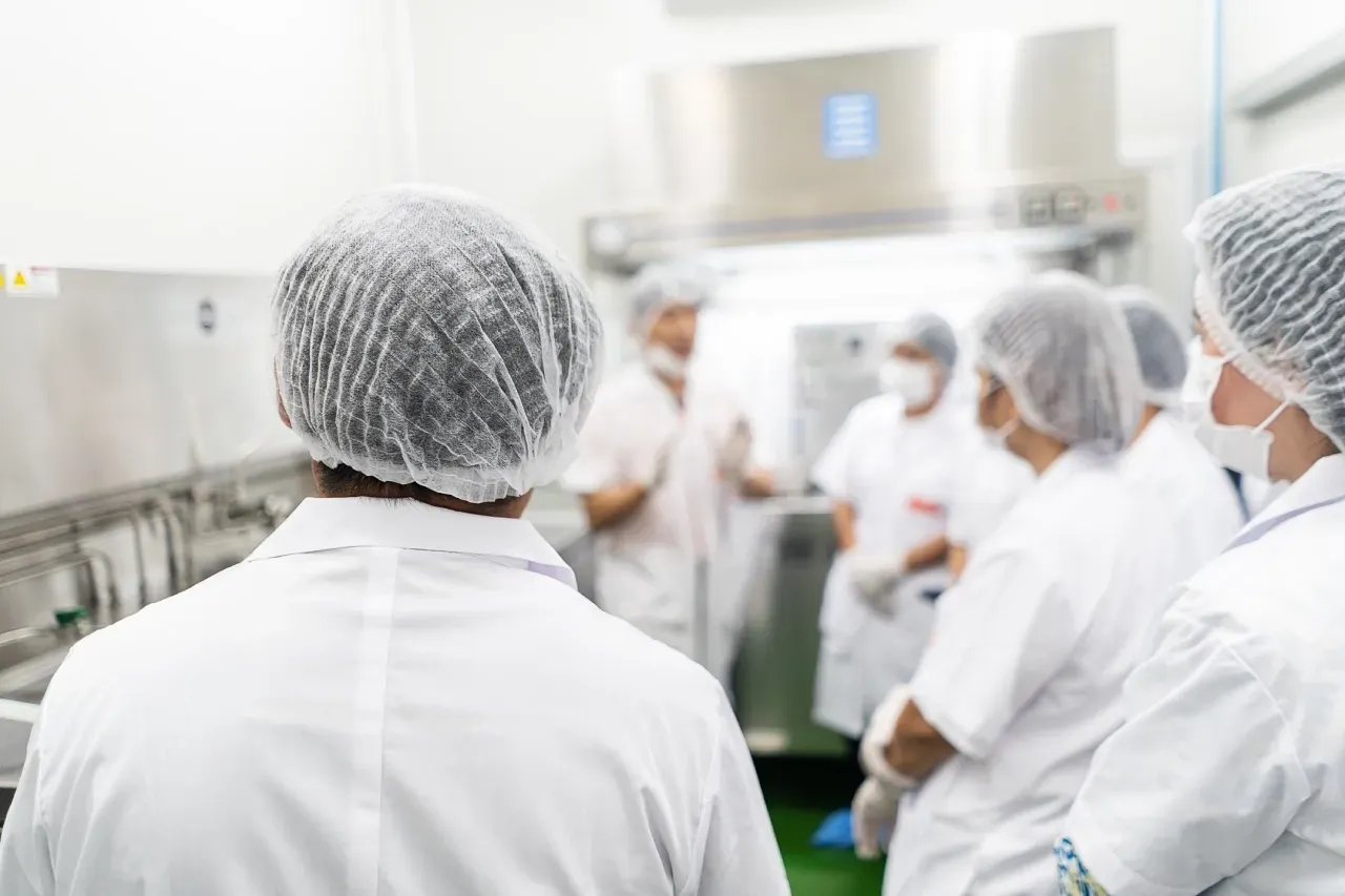 Um grupo de pessoas vestindo jalecos brancos e chapéus de plástico estão em uma cozinha.