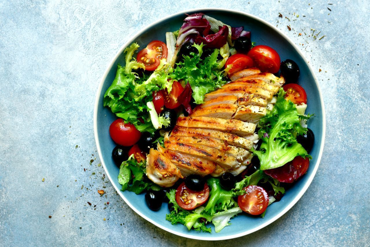 Um prato de comida com frango, tomates, azeitonas e alface sobre uma mesa.