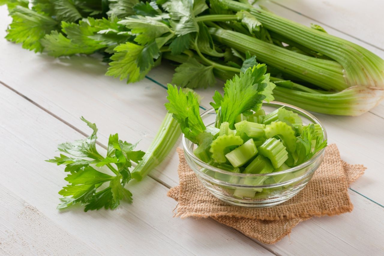 Salsão fresco com talos e folhas verdes, parte inteira e pedaços cortados em uma tigela sobre superfície clara.