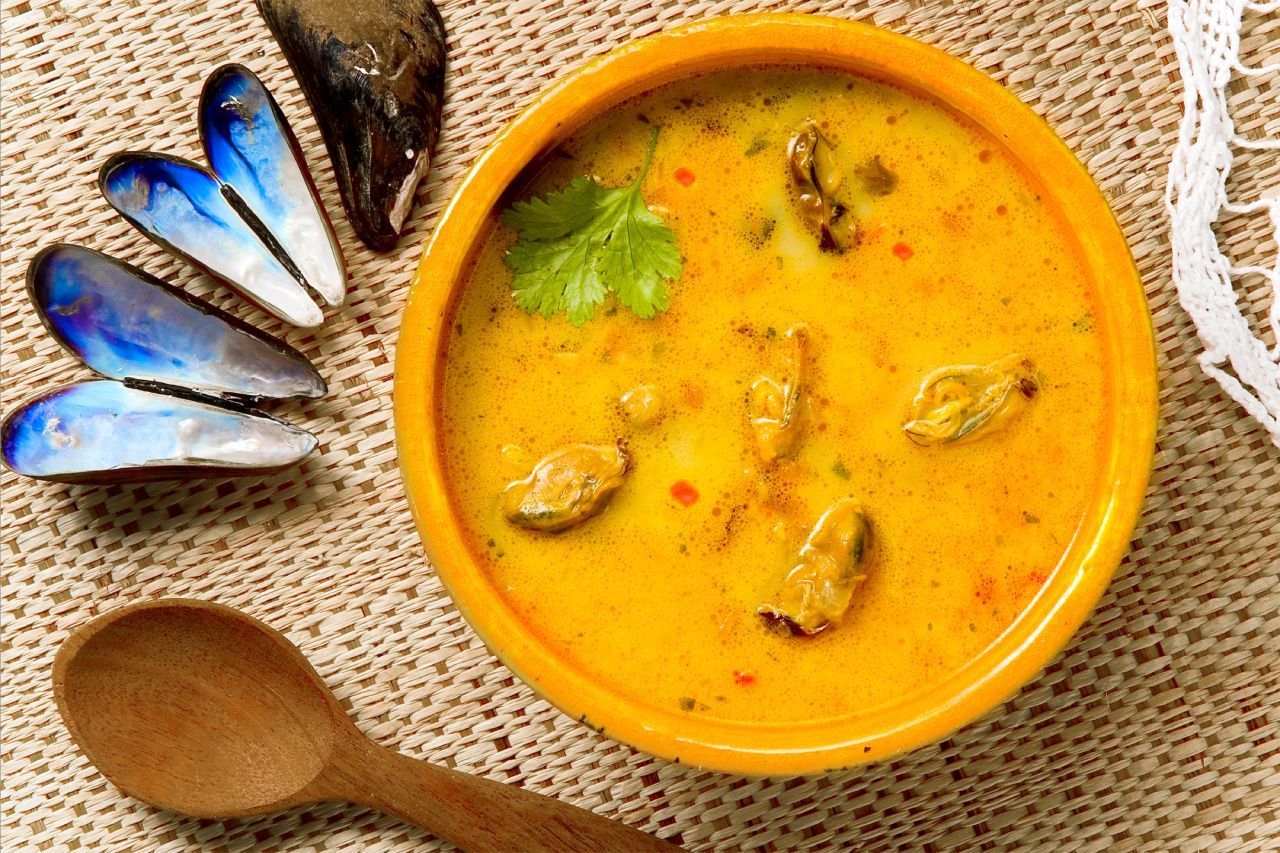 Uma tigela de sopa com mexilhões e uma colher de pau sobre uma mesa.