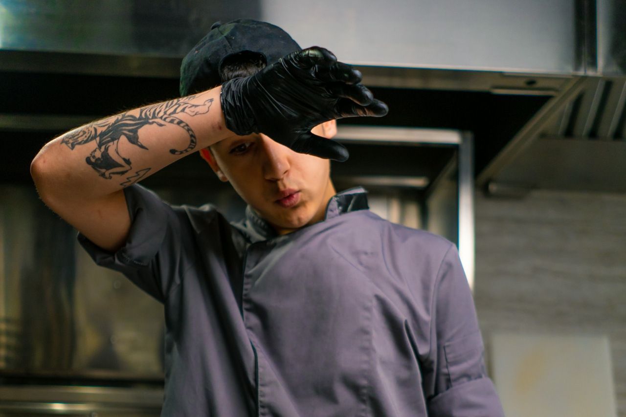 Um homem com uma tatuagem no braço está cobrindo o rosto com a mão em uma cozinha.