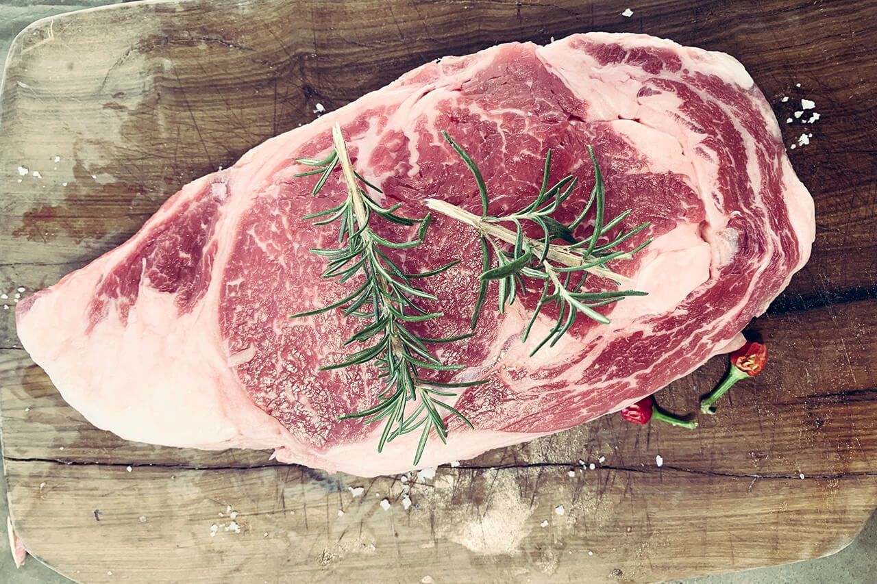 Um pedaço de bife cru com alecrim em uma tábua de corte de madeira.