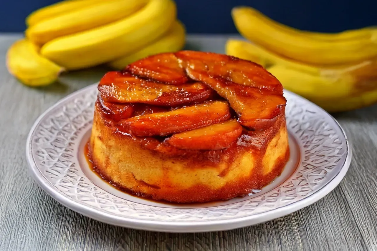 Um bolo de banana de cabeça para baixo em um prato branco com bananas ao fundo.