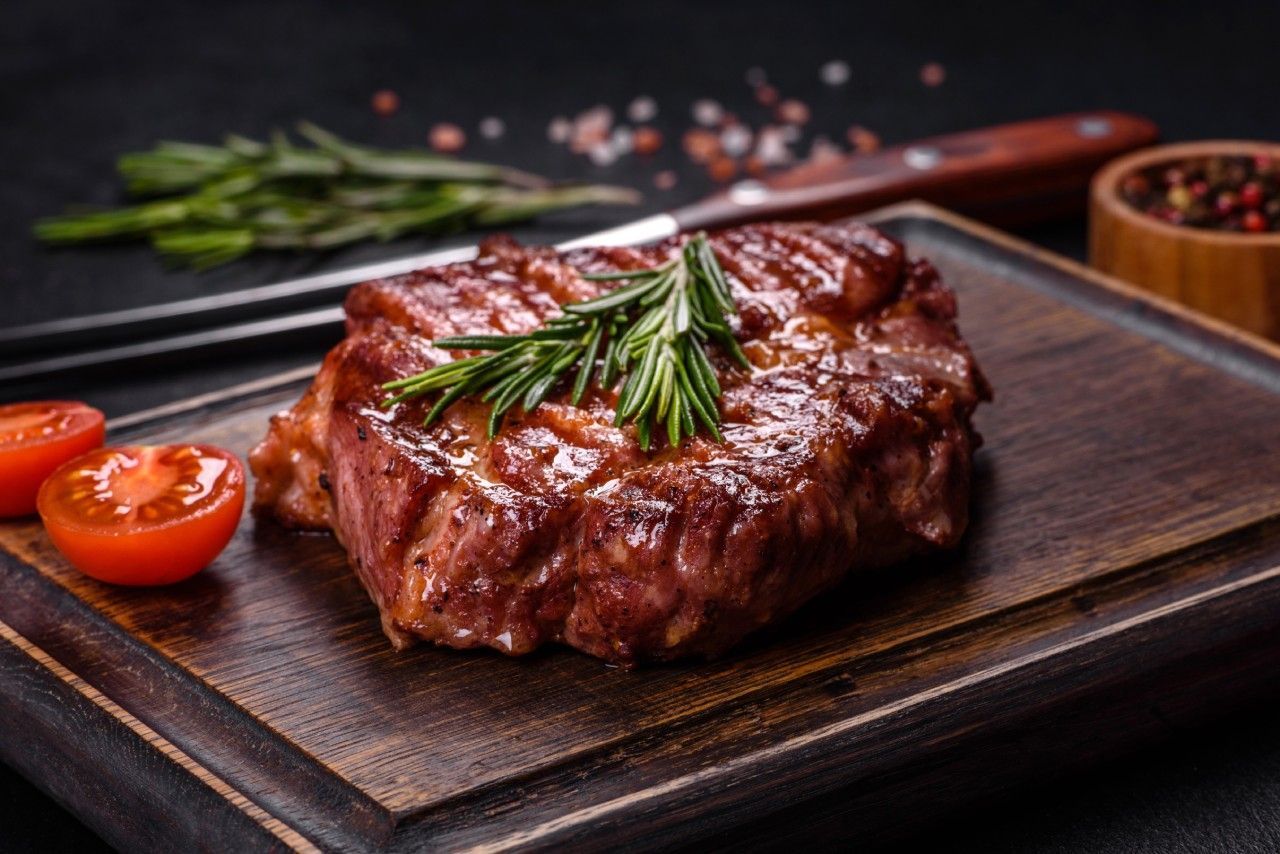 Um bife está sobre uma tábua de madeira com tomates e alecrim.