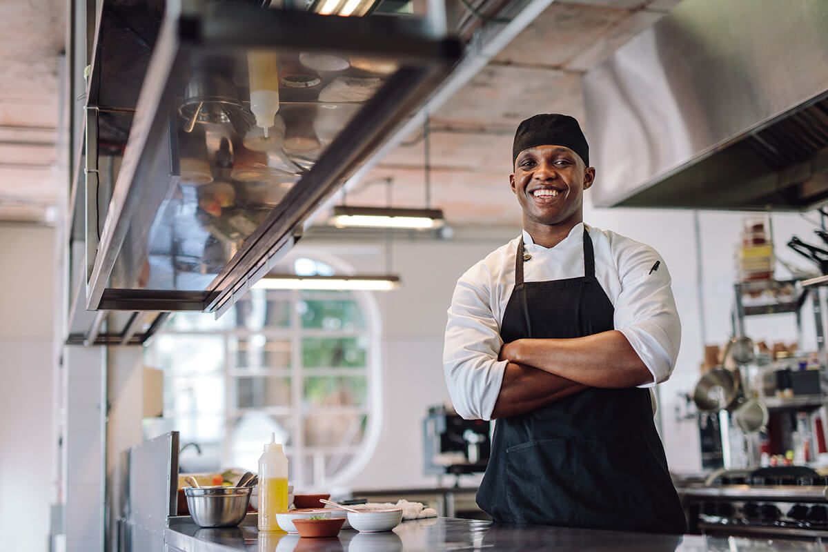 Um chef está de pé em uma cozinha com os braços cruzados e sorrindo.