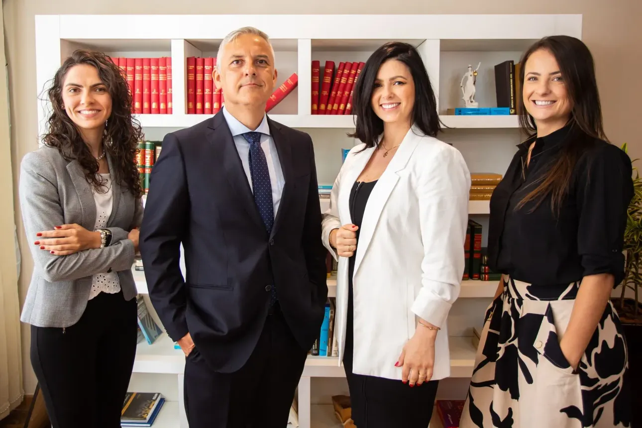 Quatro profissionais em pé em um escritório, posando em frente a estantes de livros e objetos decorativos.