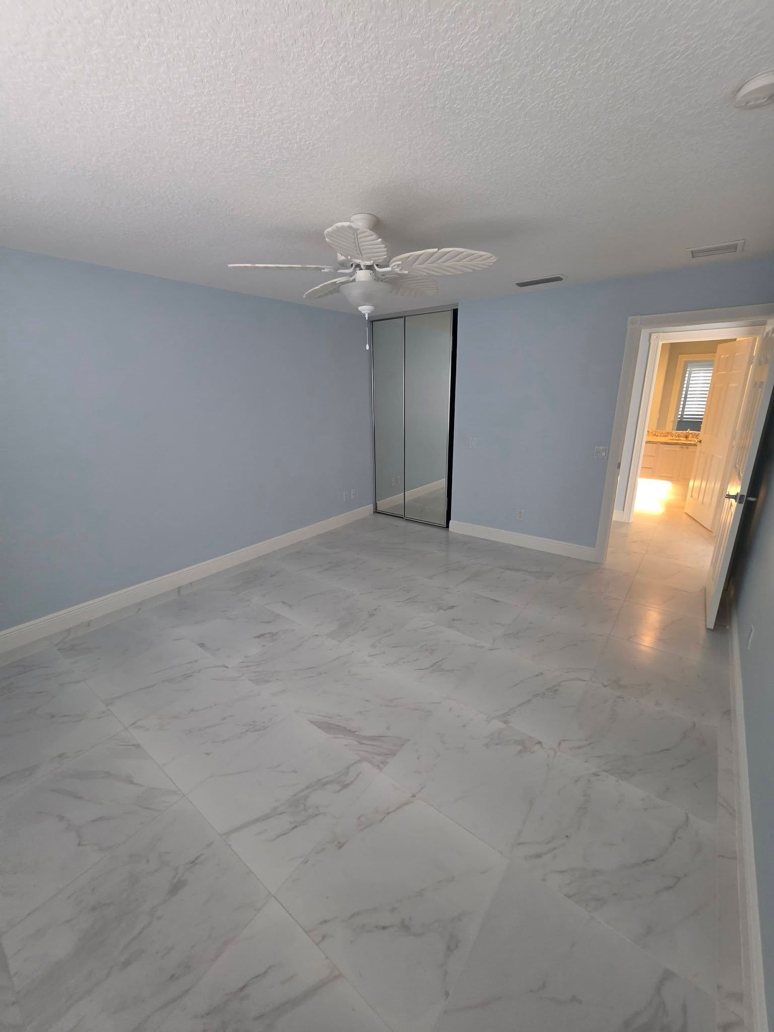 Empty light-blue bedroom with white marble-pattern floor, ceiling fan, mirrored closet, and doorway to hall.