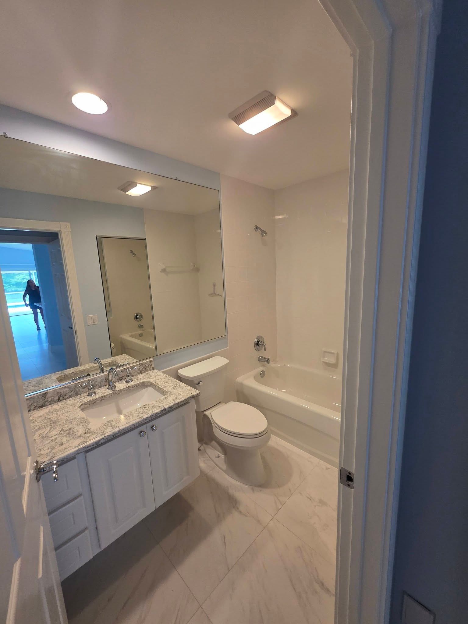 Bright bathroom with granite vanity, large mirror, toilet, and tub under ceiling lights.
