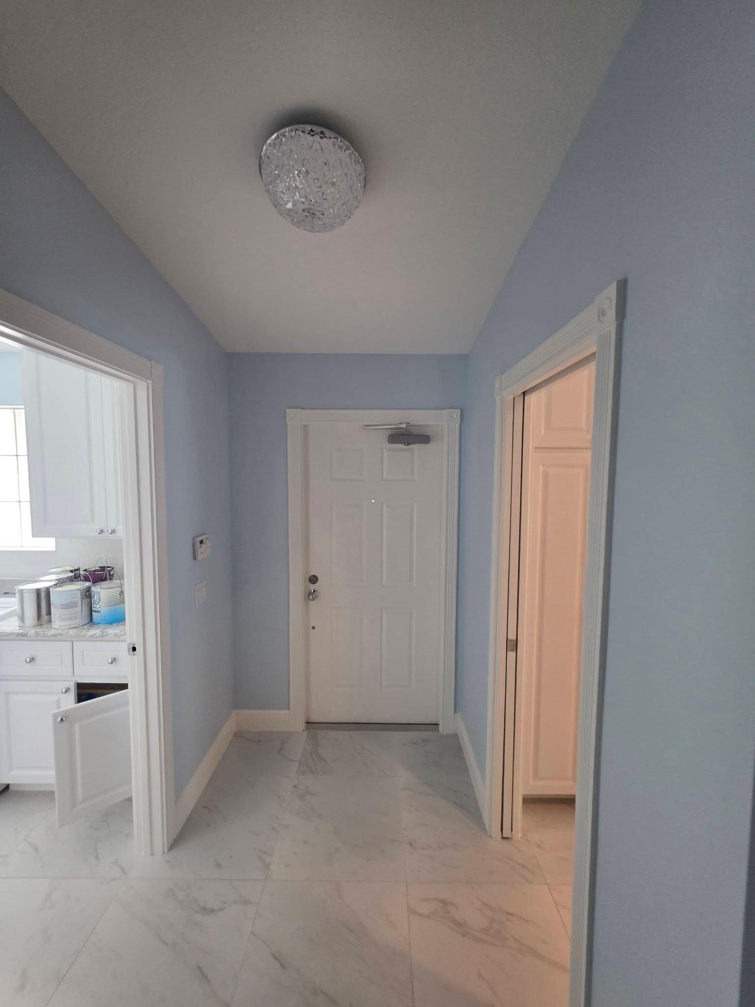 Narrow hallway with light blue walls, white door, marble floor, and kitchen visible on the left.