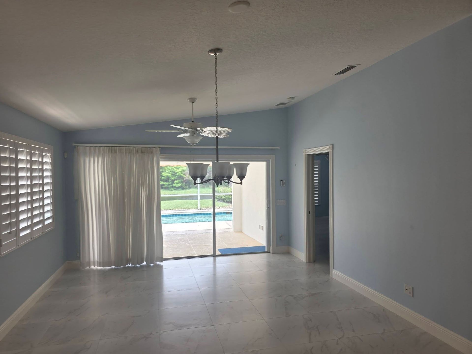 Empty living room with light blue walls, tile floor, ceiling fan, sliding glass doors, and a view of the backyard.