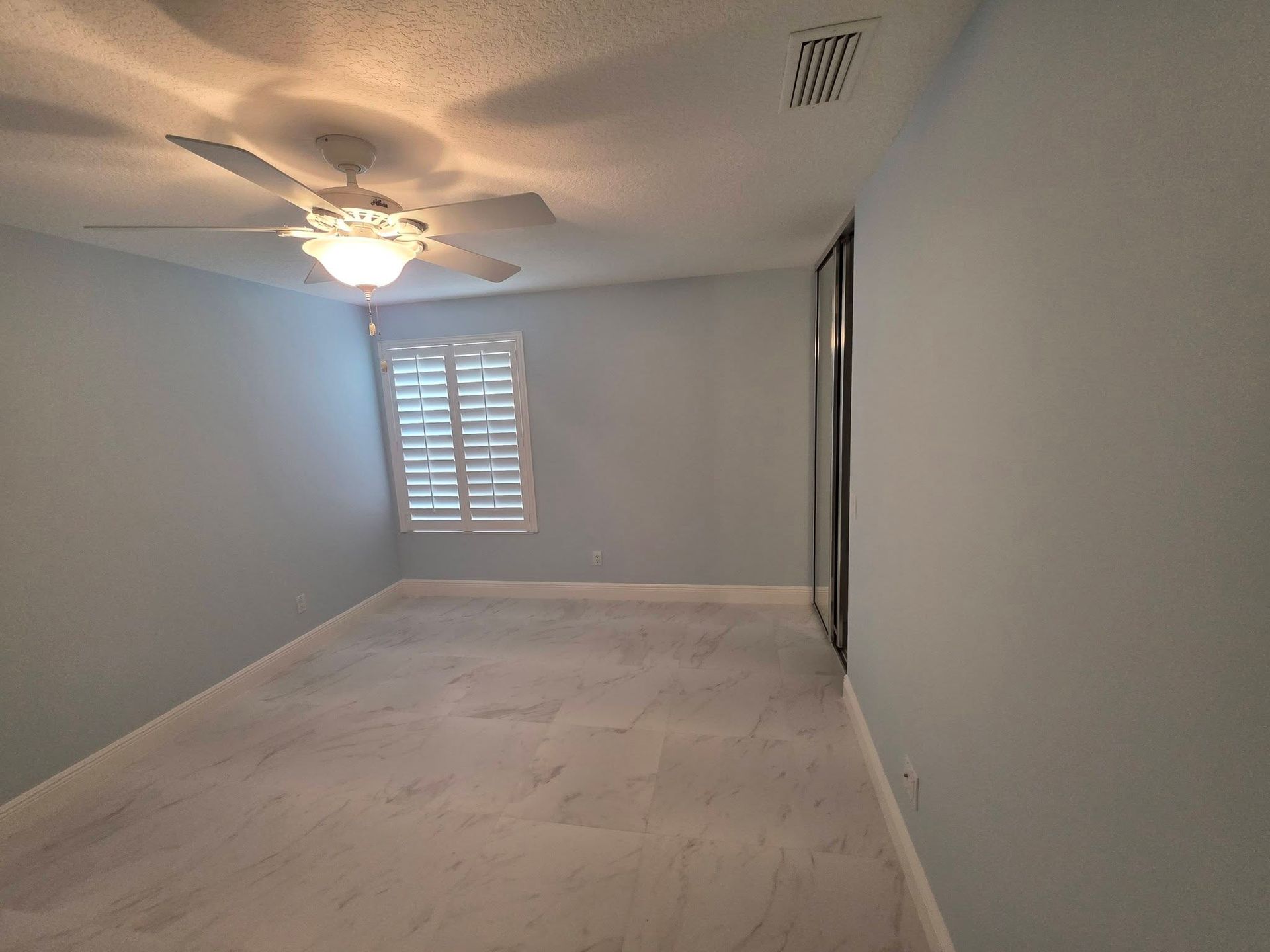 Empty bedroom with light gray walls, ceiling fan, and window shutters, viewed from the doorway