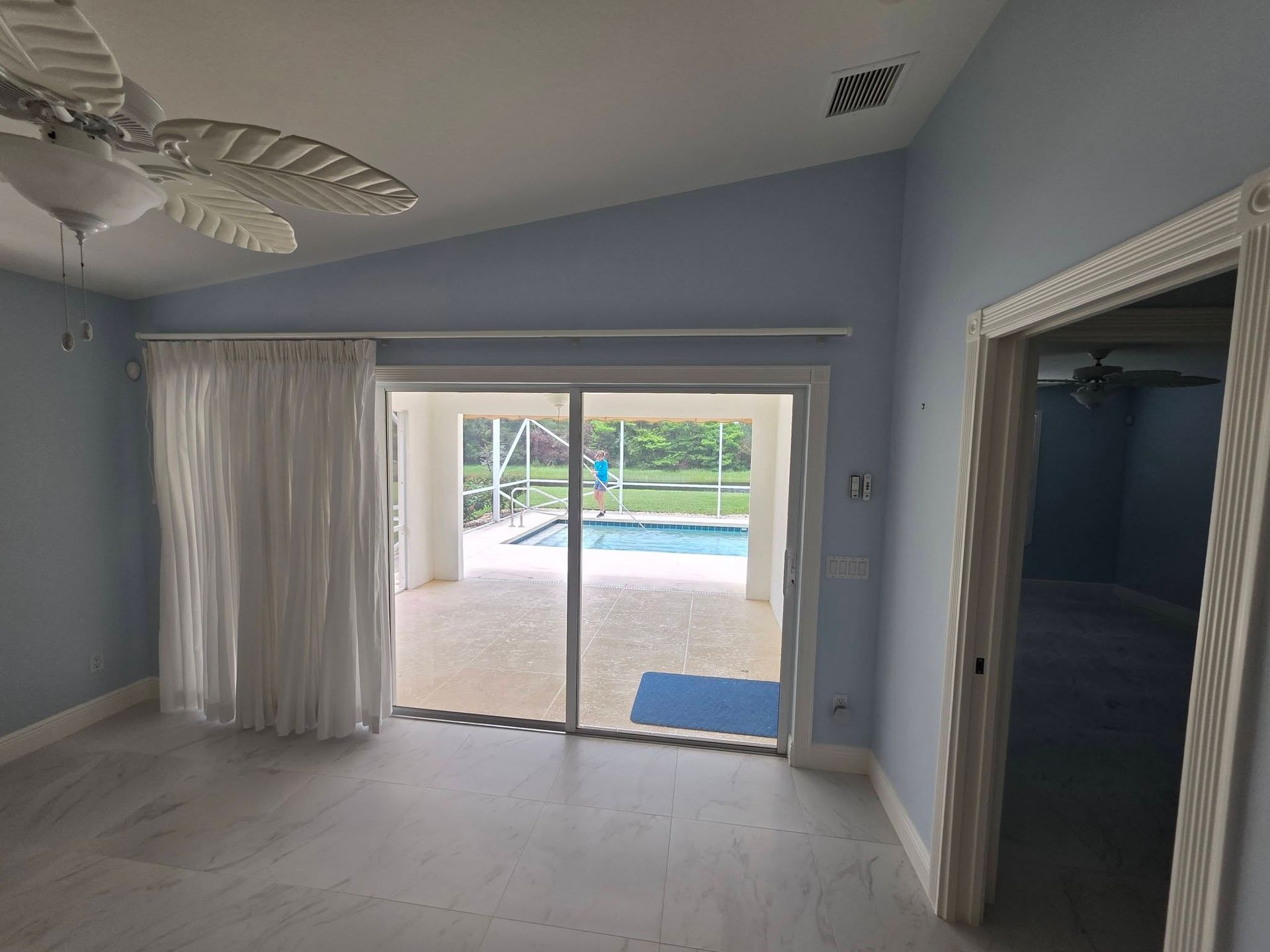 Empty living room with white curtains, sliding glass doors, and a view of a backyard pool