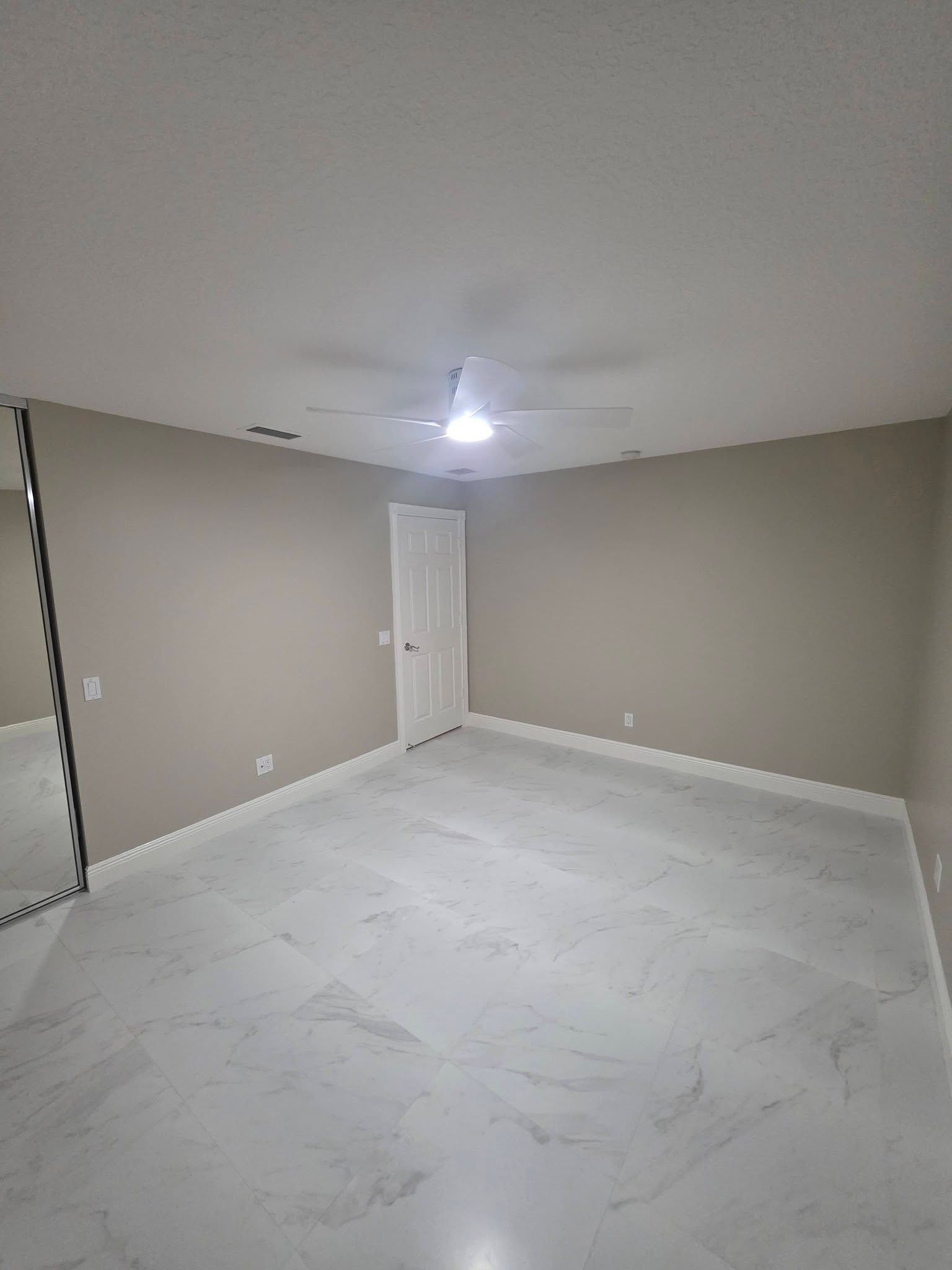 Empty beige room with white floor, single light bulb, and closed white door under a low ceiling.