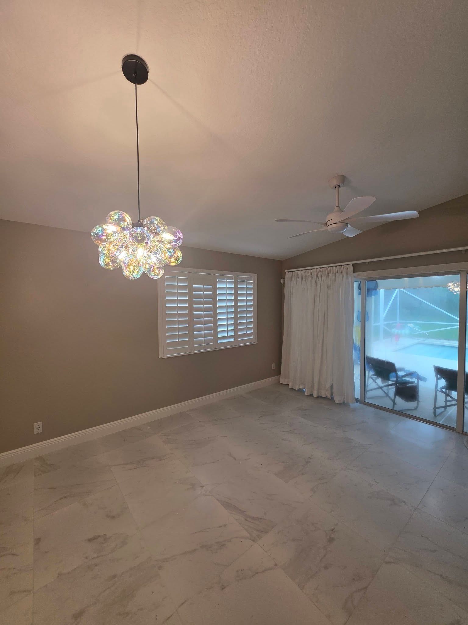 Empty living room with chandelier, window shutters, ceiling fan, and sliding glass doors to a patio.