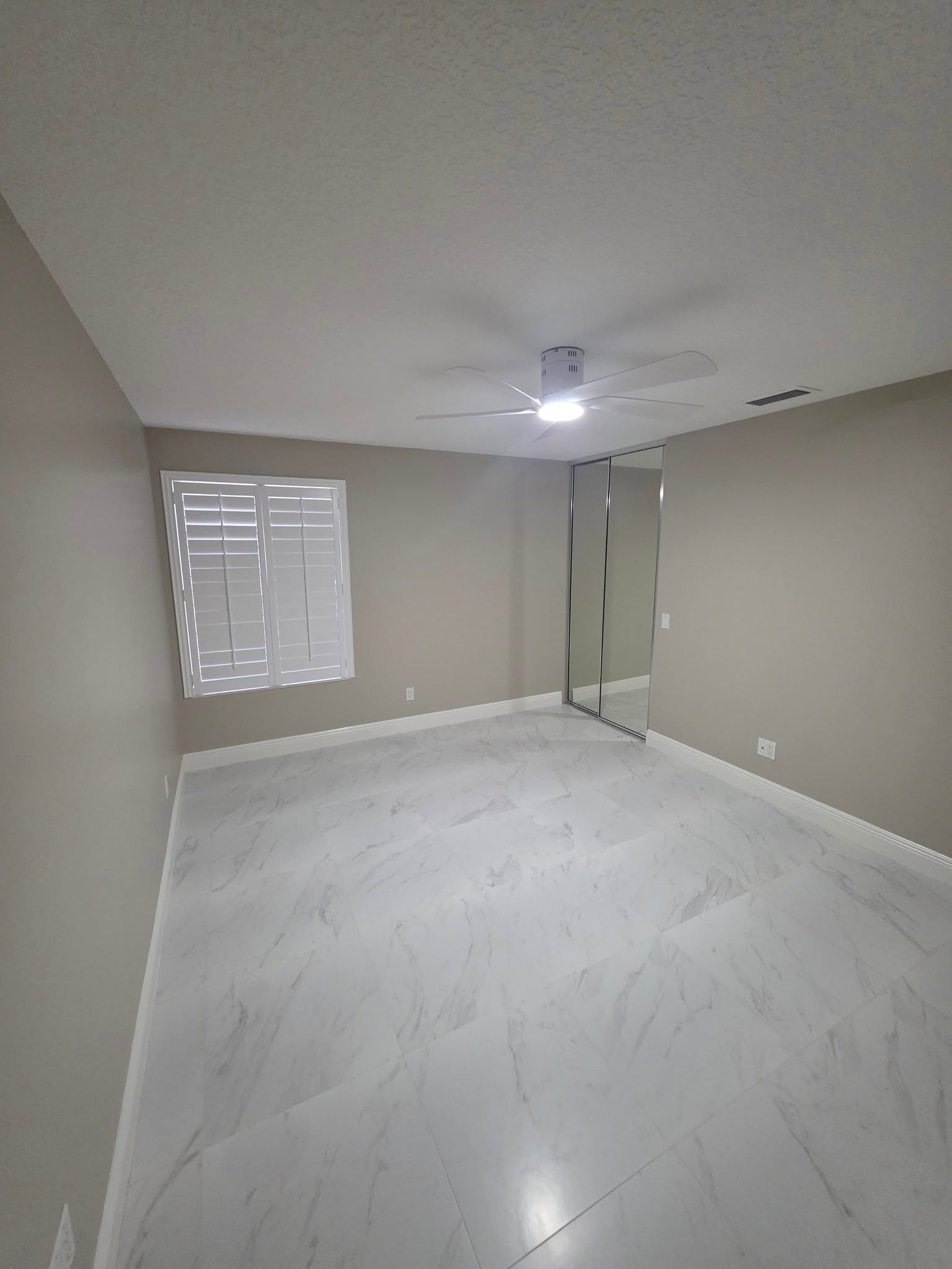 Empty beige bedroom with white marble floors, mirrored closet doors, and a ceiling fan.
