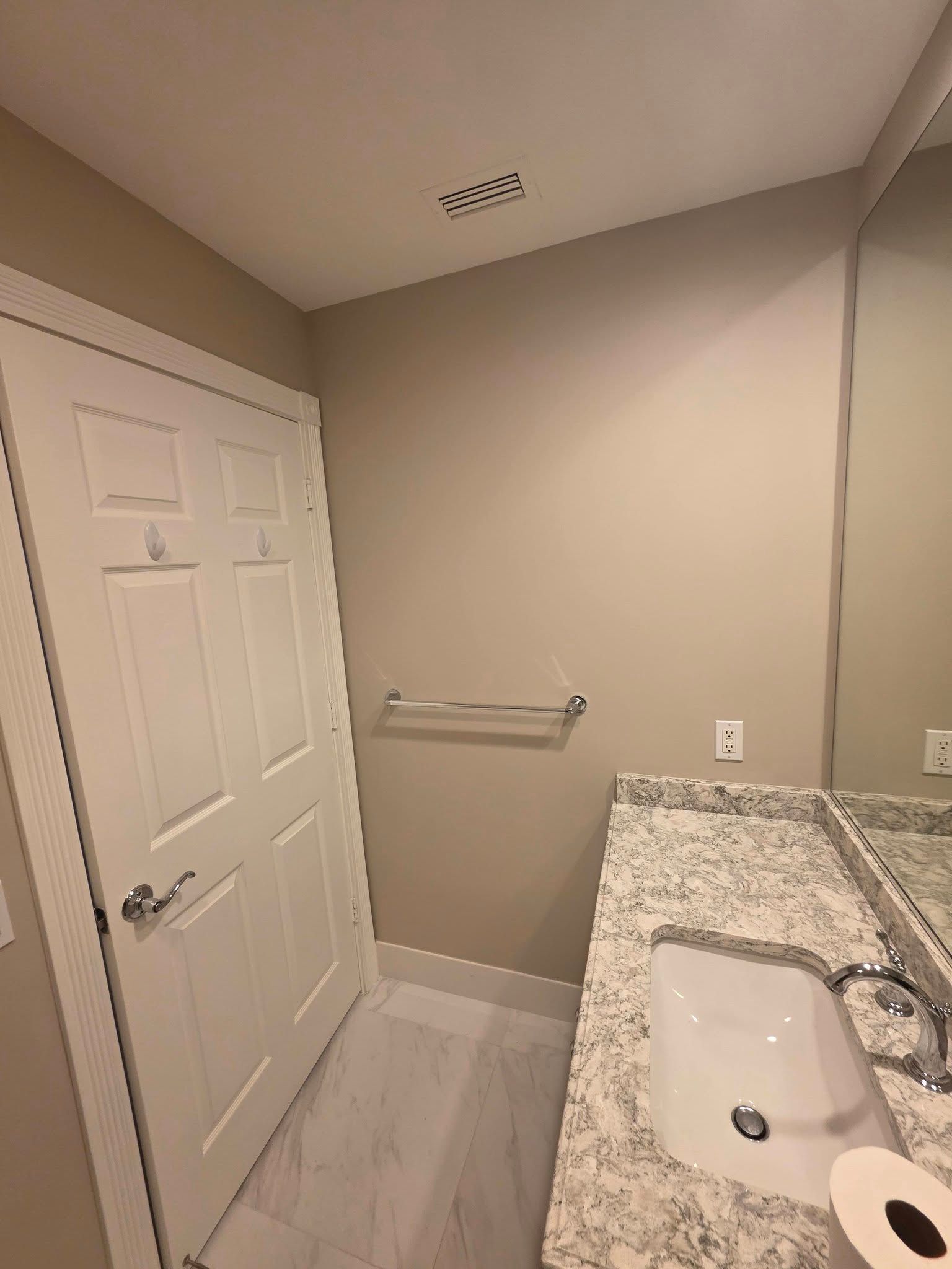 Small bathroom with white door, granite vanity, wall mirror, towel rack, and toilet paper roll.