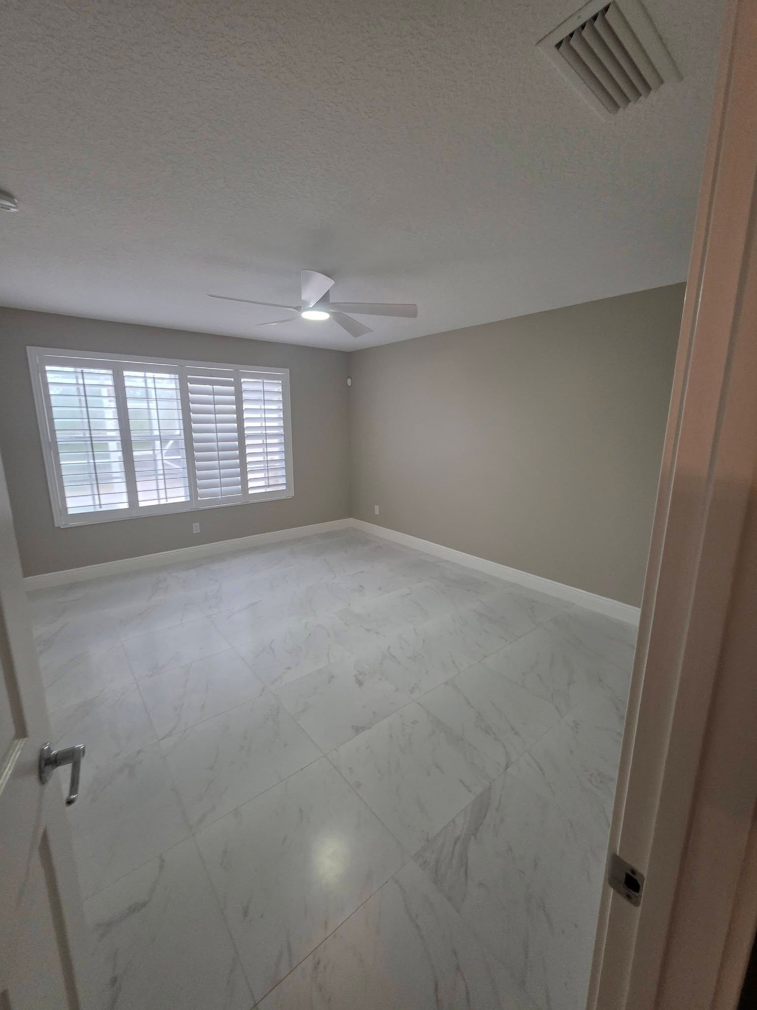 Empty beige room with white tile floor, ceiling fan, and window blinds.