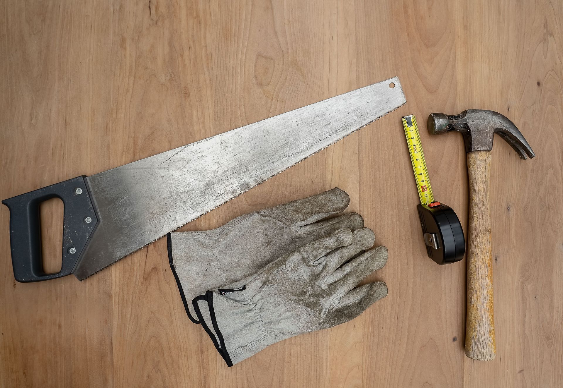 Handsaw, work gloves, hammer, and tape measure laid out on a wooden surface.