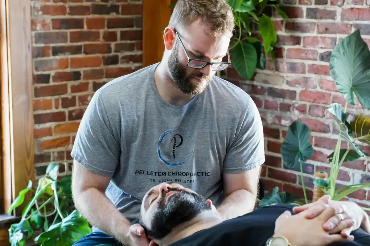 Bearded man in gray T-shirt gently holds a person reclining indoors among brick walls and plants