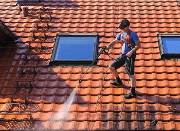 Man power washing a red tiled roof, wearing a safety harness.