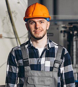 Man in orange hard hat, plaid shirt, and overalls smiling in a workshop setting.