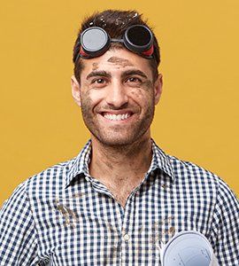Man wearing goggles, covered in dirt, smiling broadly. Blue and white plaid shirt. Yellow background.