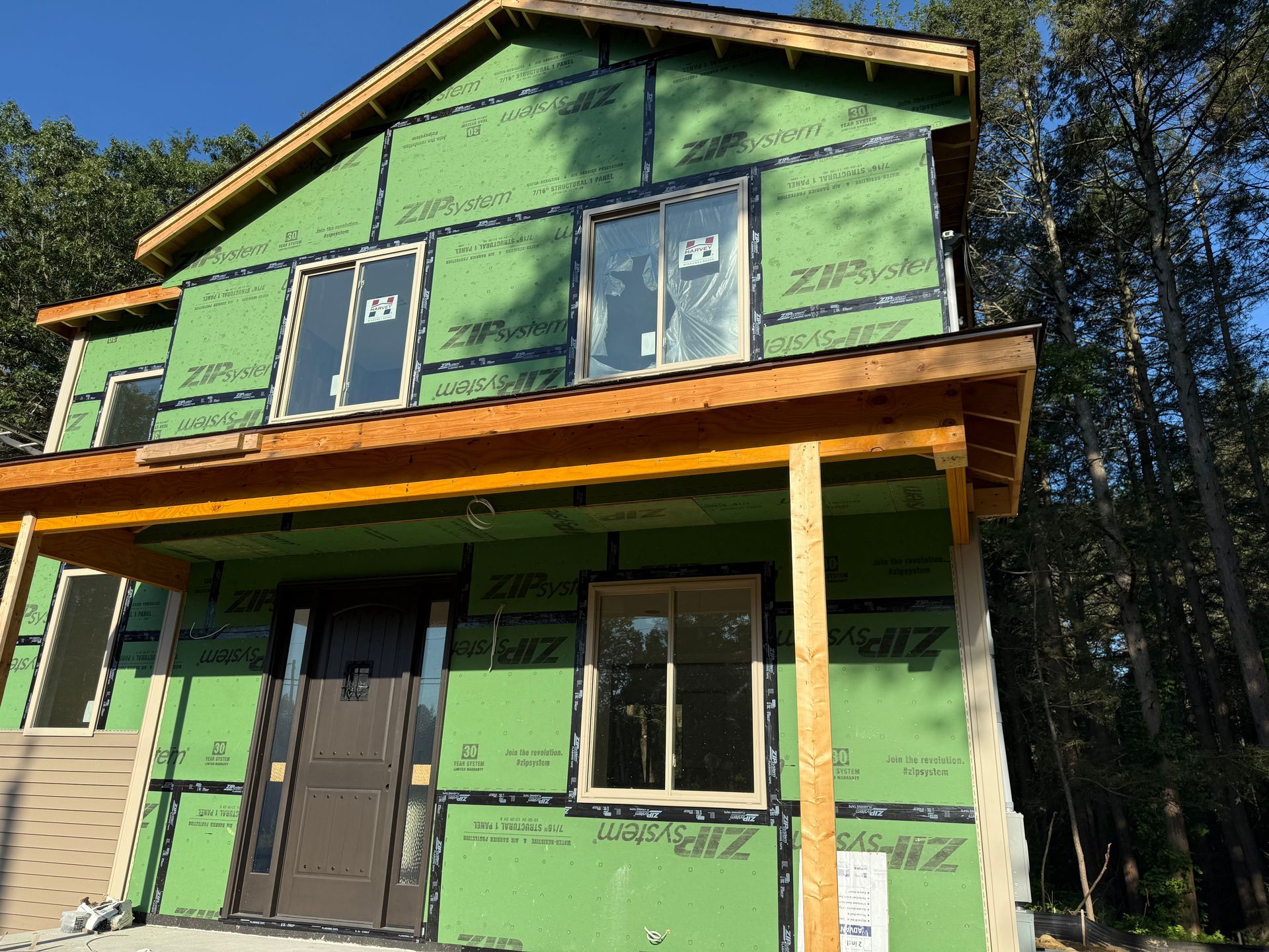 Two-story house under construction, green sheathing, windows, brown door and wood frame porch, set in wooded area.