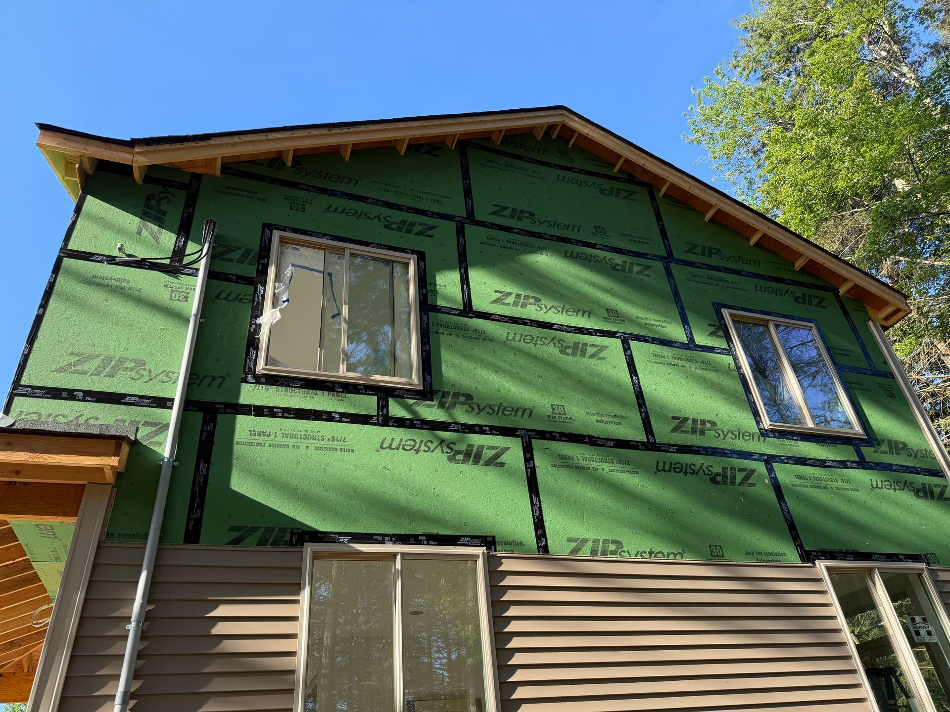 Two-story house under construction, with green sheathing and brown siding, windows, and clear blue sky.