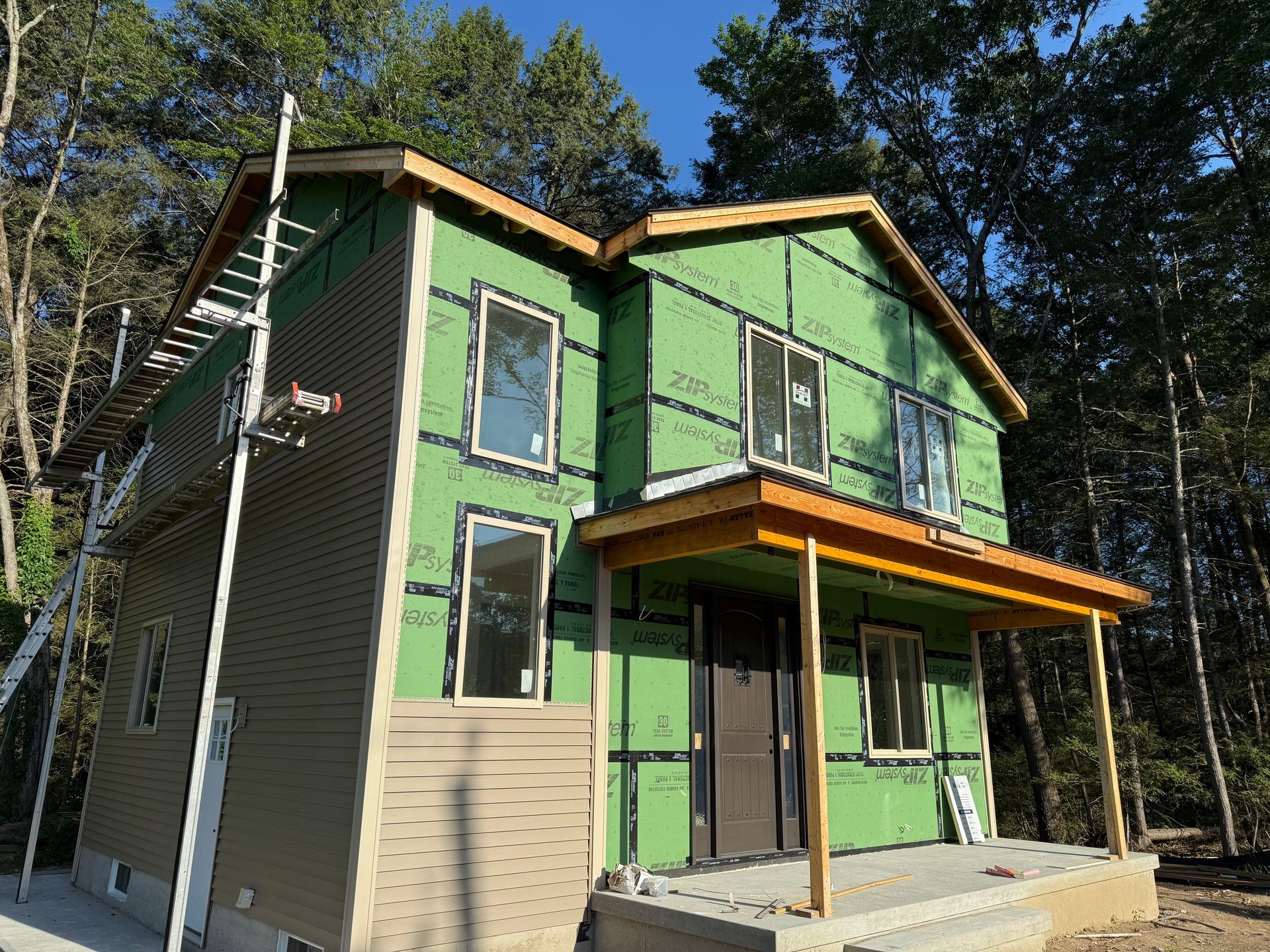 Two-story house under construction, green sheathing, brown siding, scaffolding, forest background.