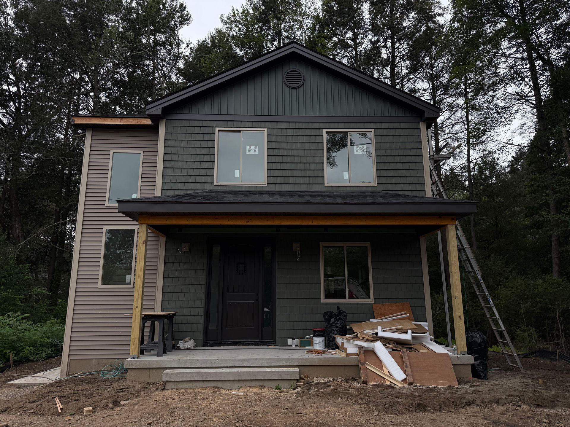 Two-story house with dark green siding, covered porch, and construction materials scattered on the porch.