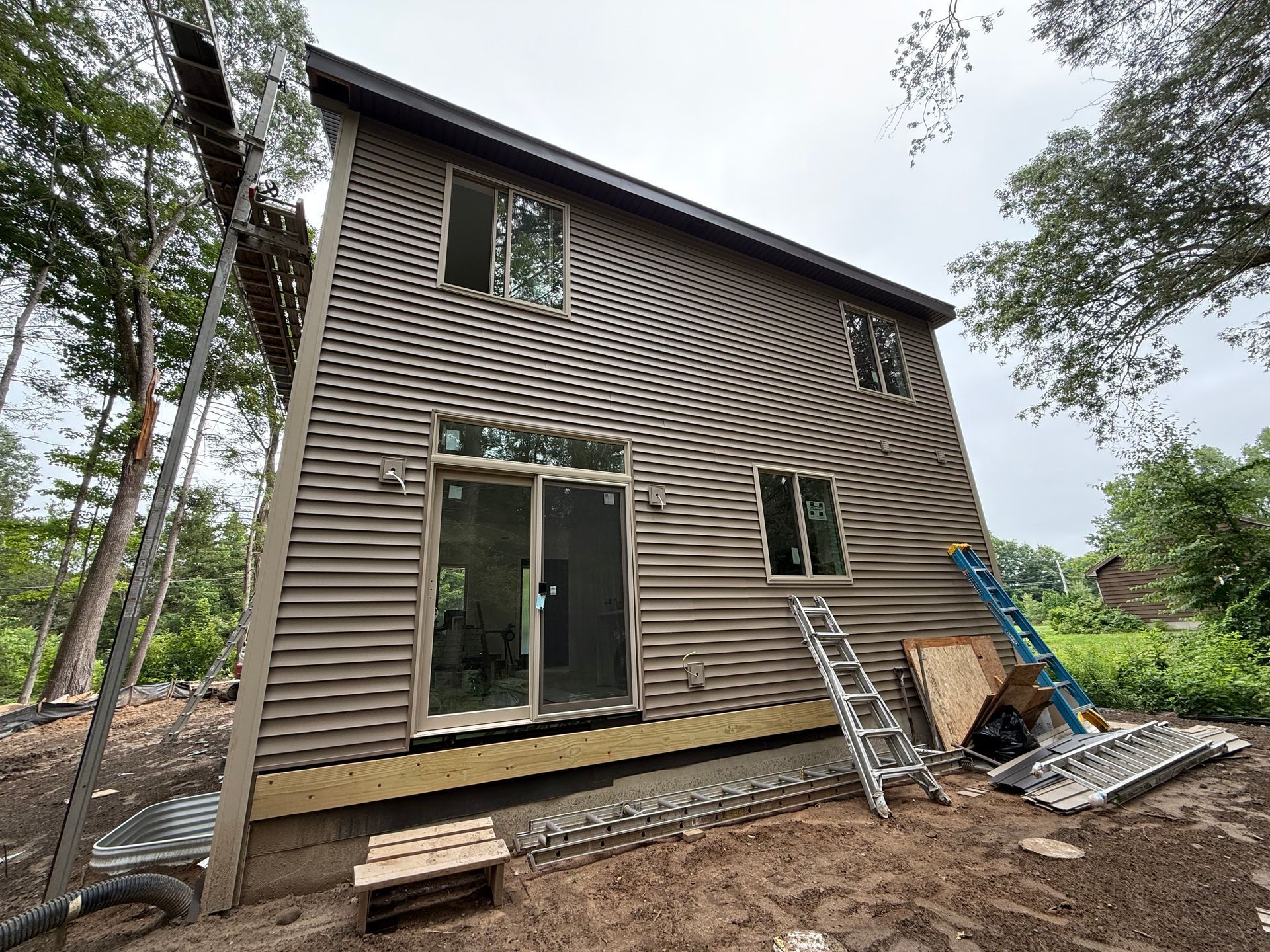 Two-story house with brown siding, sliding glass door, and a ladder leaning against it. Cloudy outdoor setting.